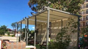 Un espace extérieur abrité, doté d’une structure en forme de pergola, entouré de verdure et bordé d’un mur en briques.