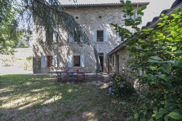 A stone building with a courtyard, a table with chairs, and a garden with purple flowers.
