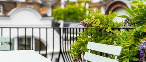 A balcony with a white table and a light green chair, surrounded by lush greenery and purple flowers.