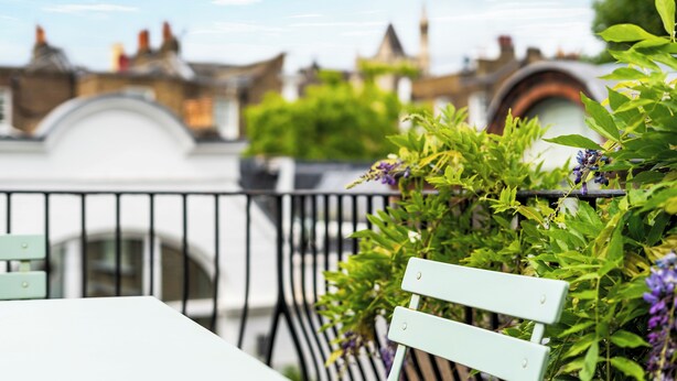 A balcony with a white table and a light green chair, surrounded by lush green plants and purple flowers.
