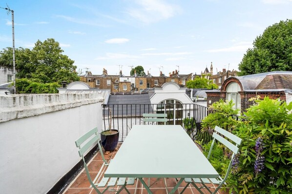 A rooftop terrace with a table and chairs, surrounded by greenery and overlooking a residential area.