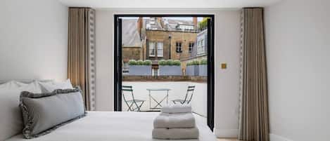 A modern bedroom with a large bed, a small table and chairs, and a view of a courtyard through an open door.