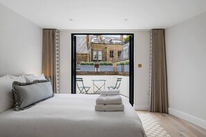 A modern bedroom with a large bed, a small table and chairs, and a view of a courtyard through an open door. - Elegant split level High St Ken Apt with Terrace (London)