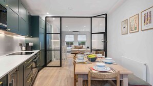 A modern kitchen with a dining area, featuring a wooden table set for four, a kitchen island, and a view into the living room. - Elegant split level High St Ken Apt with Terrace (London)