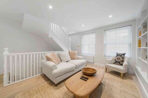 Un salon moderne avec un escalier blanc, un canapé, un fauteuil à rayures, une table basse en bois et une grande fenêtre.