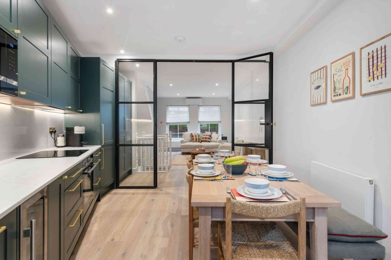 A modern kitchen with a dining area, featuring a central island, wooden flooring, and a view to a living room.