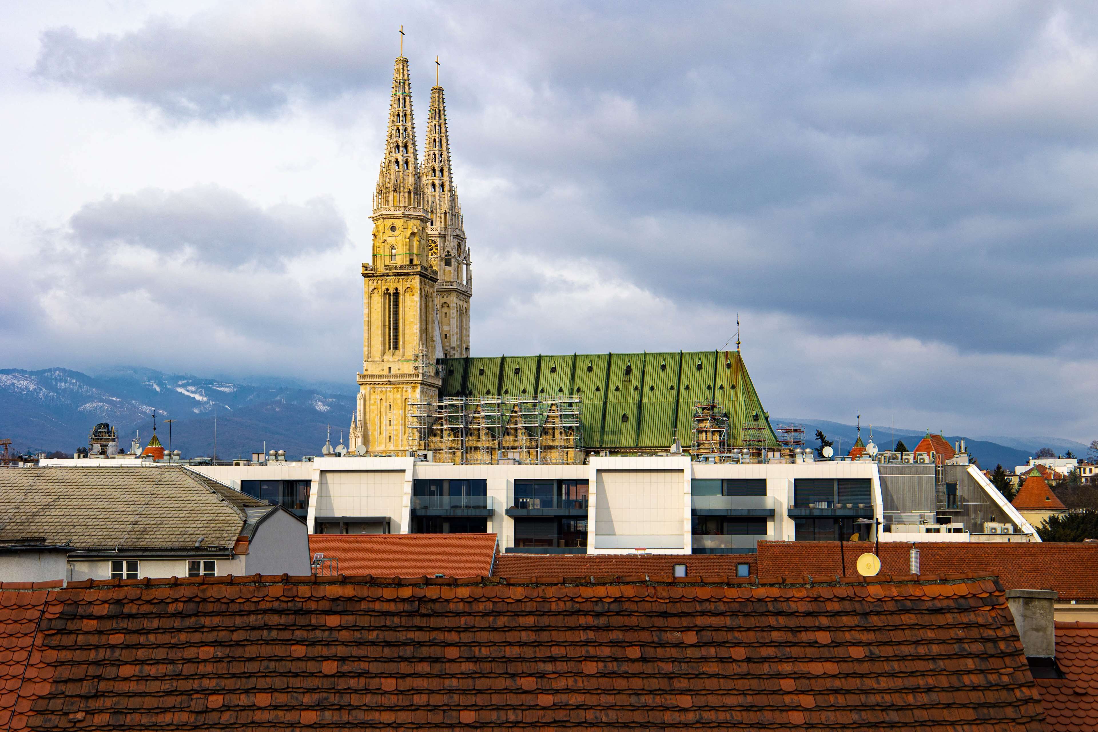 Uma catedral histórica com uma torre alta, edifícios modernos com andaimes e um cenário montanhoso ao fundo.