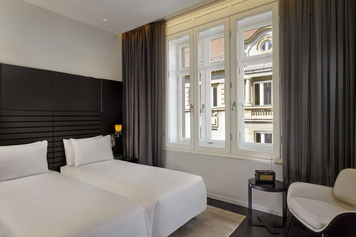 Une chambre d’hôtel avec deux lits, une petite table avec une radio, une chaise et une vue sur un immeuble à travers de grandes fenêtres.