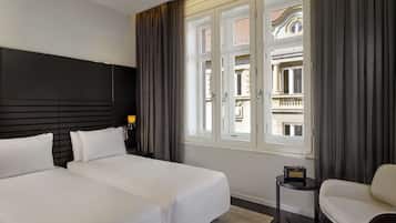 Une chambre d’hôtel avec deux lits, une petite table avec une radio, une chaise et une vue sur un immeuble à travers de grandes fenêtres.