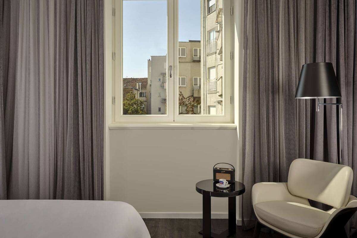 Une chambre d’hôtel avec un lit, une chaise, une petite table avec une radio et une fenêtre avec des rideaux.