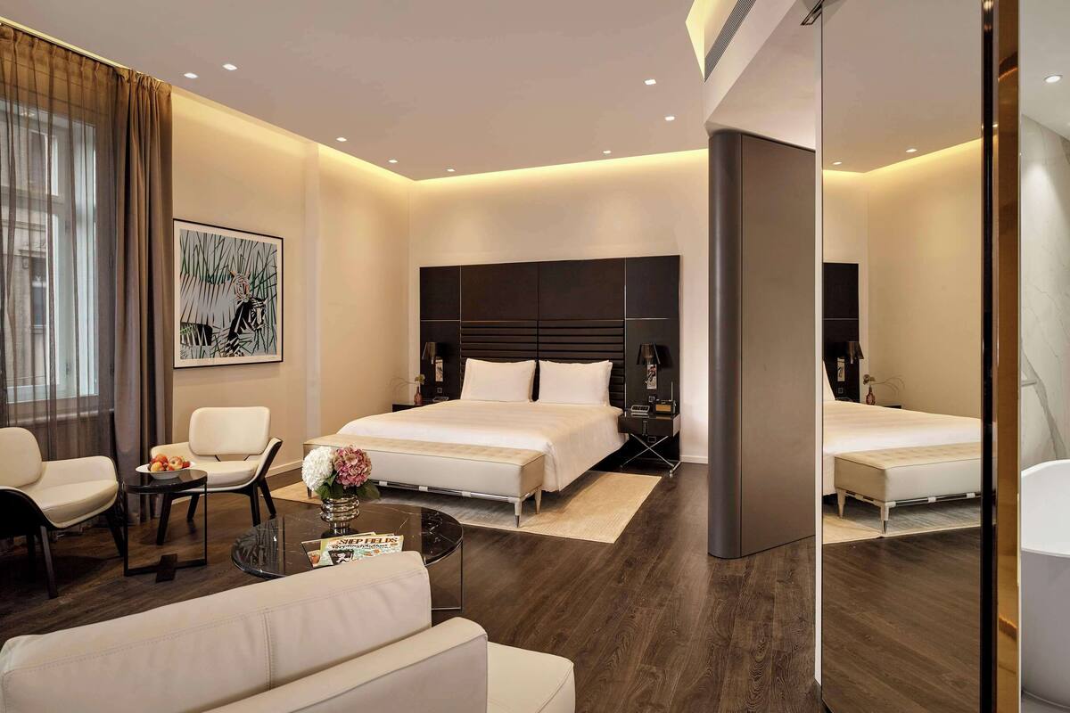 Une chambre d’hôtel moderne avec deux lits, un coin salon avec un canapé et des fauteuils, une petite table agrémentée d’un vase de fleurs, une grande œuvre d’art au mur et une salle de bain séparée avec une baignoire.