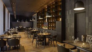 Een modern restaurantinterieur met houten vloeren, donkerbruine tafels en stoelen, hanglampen en een wand met planken waarop flessen en glazen staan.