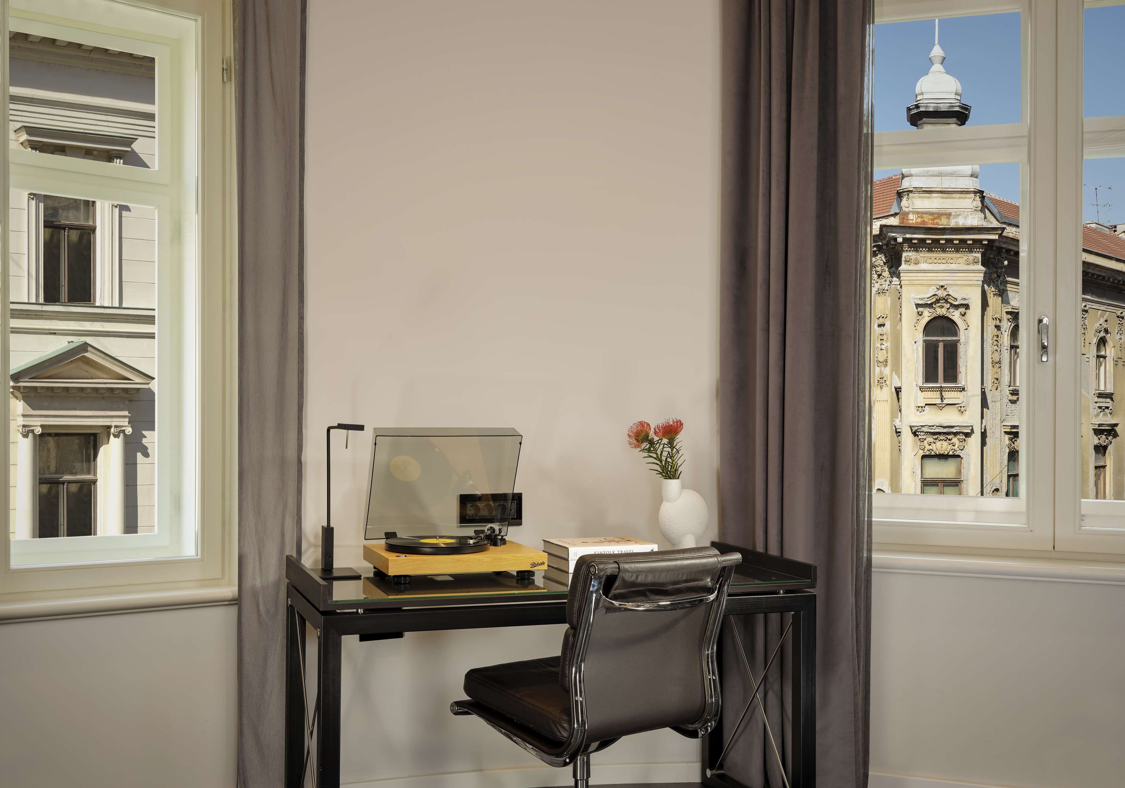 A room with a desk, a chair, a computer, a vase with flowers, and a view of a historic building.