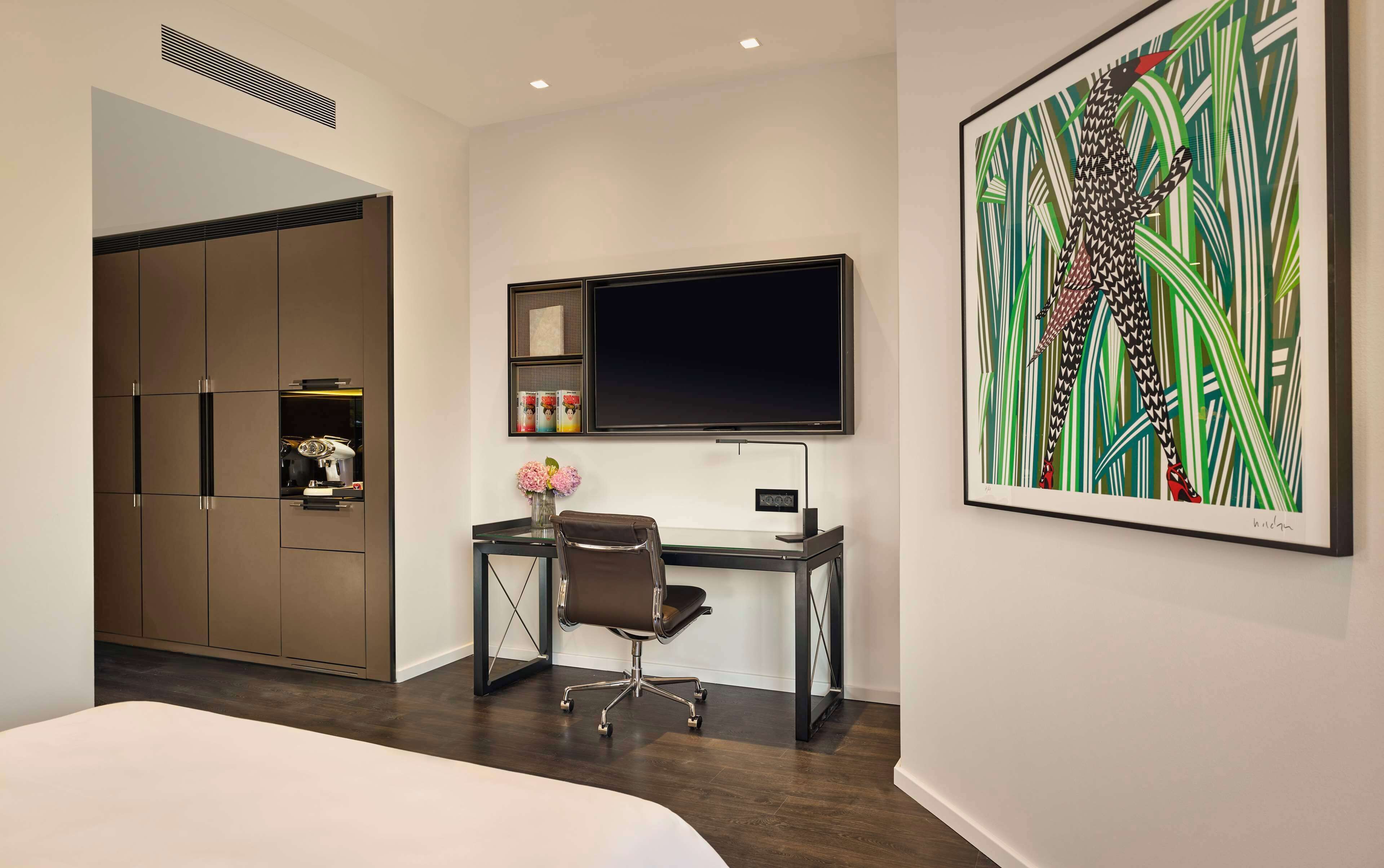 Une chambre d’hôtel moderne équipée d’un lit, d’un bureau avec un ordinateur, d’une télévision et d’un grand tableau abstrait.