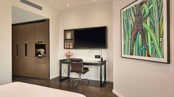 A modern hotel room with a bed, a desk with a computer, a TV, and a large abstract painting.