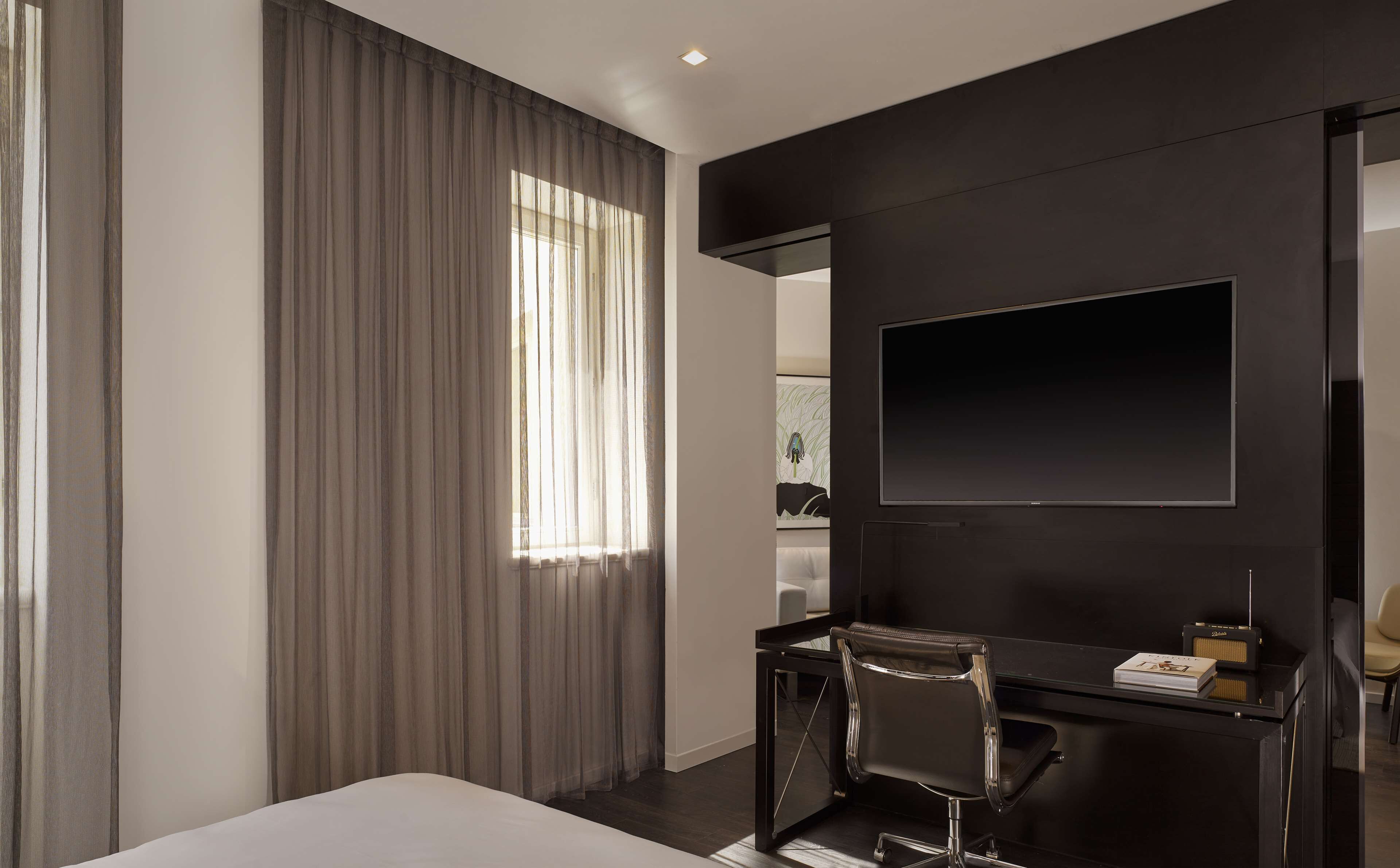 Ein modernes Hotelzimmer mit einem großen Bett, einem Schreibtisch mit Stuhl, einem Flachbildfernseher und einem großen Fenster mit Vorhängen.