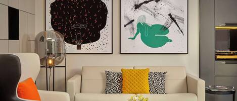 A modern living room with a sofa, armchair, and coffee table. There are two framed artworks on the wall, a floor lamp, and a small table with a vase of flowers.