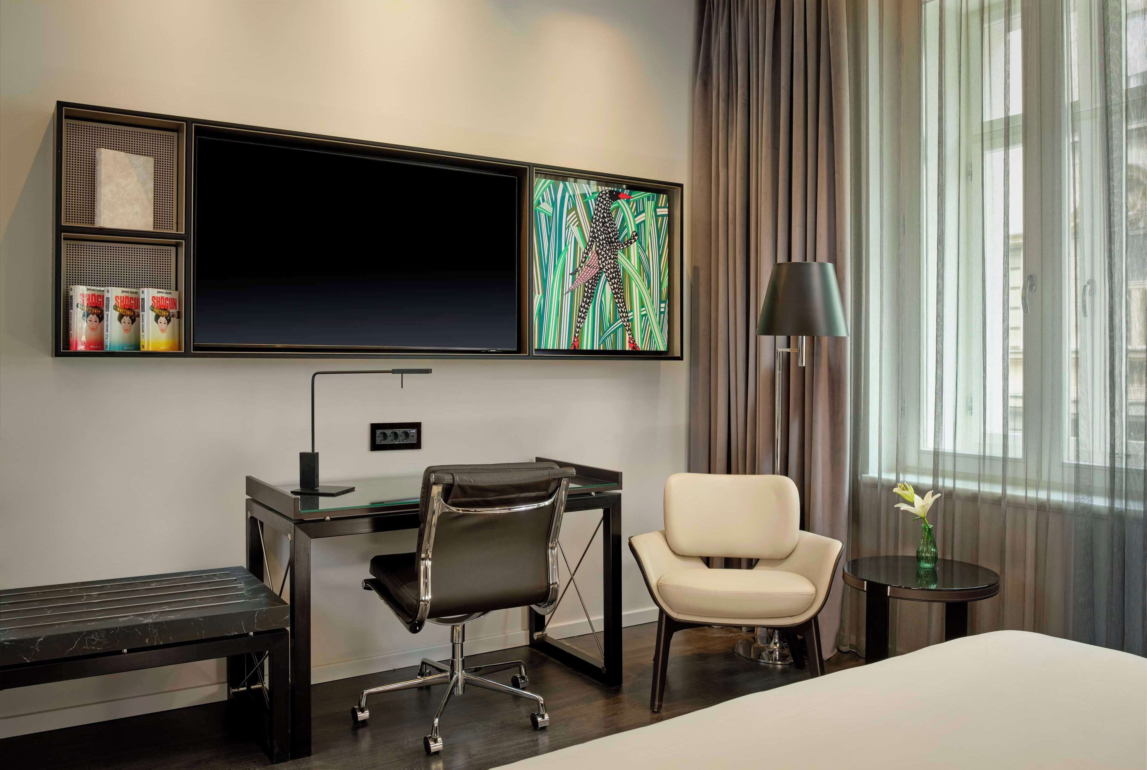 Habitación de hotel con cama, escritorio con silla, televisión, lámpara y ventana con cortinas.