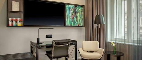 Habitación de hotel con cama, escritorio con silla, televisión, lámpara y ventana con cortinas.