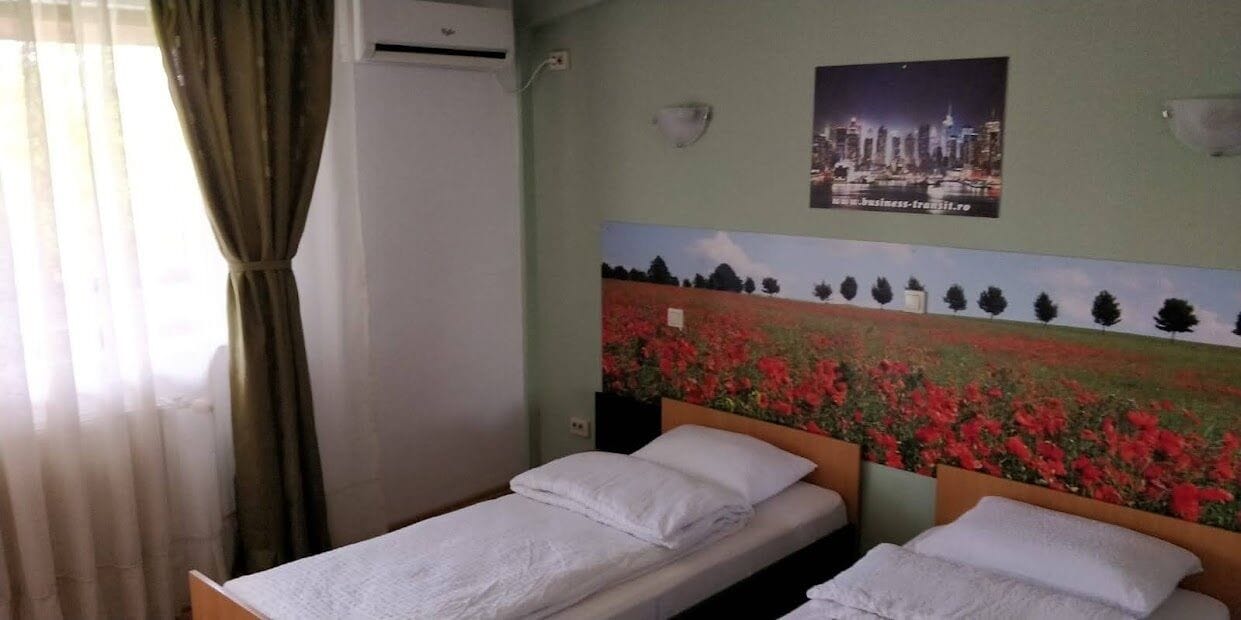 A room with two single beds, a cityscape painting on the wall, and an air conditioning unit.