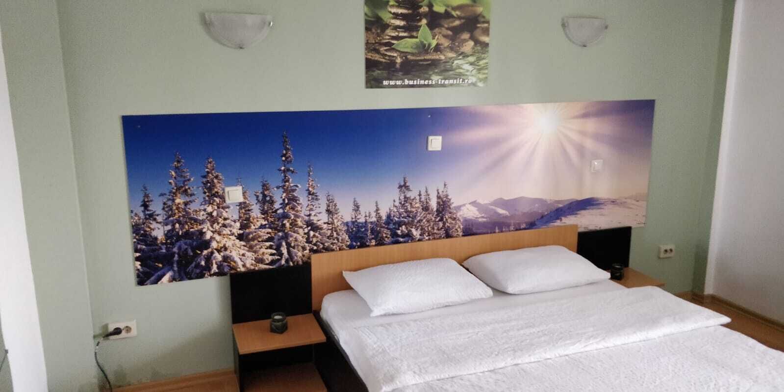 Ein Hotelzimmer mit einem großen Bett, einem Nachttisch und einem an der Wand befestigten Poster mit einer verschneiten Berglandschaft.