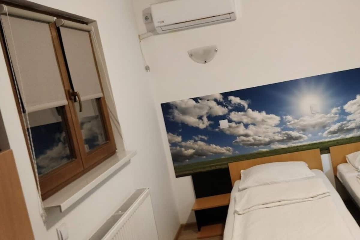 A bedroom with two beds, a window with blinds, an air conditioning unit, and a wall-mounted light fixture.