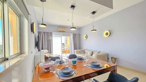 Un comedor moderno con una mesa de madera preparada para cuatro, un sofá al fondo y un balcón visible a través de la ventana.