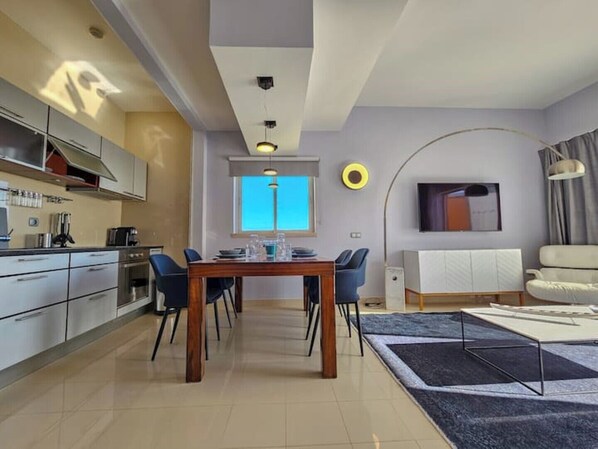 A modern kitchen with white cabinets, a dining area with a wooden table and chairs, a large window with a view, and a living area with a sofa and a TV.