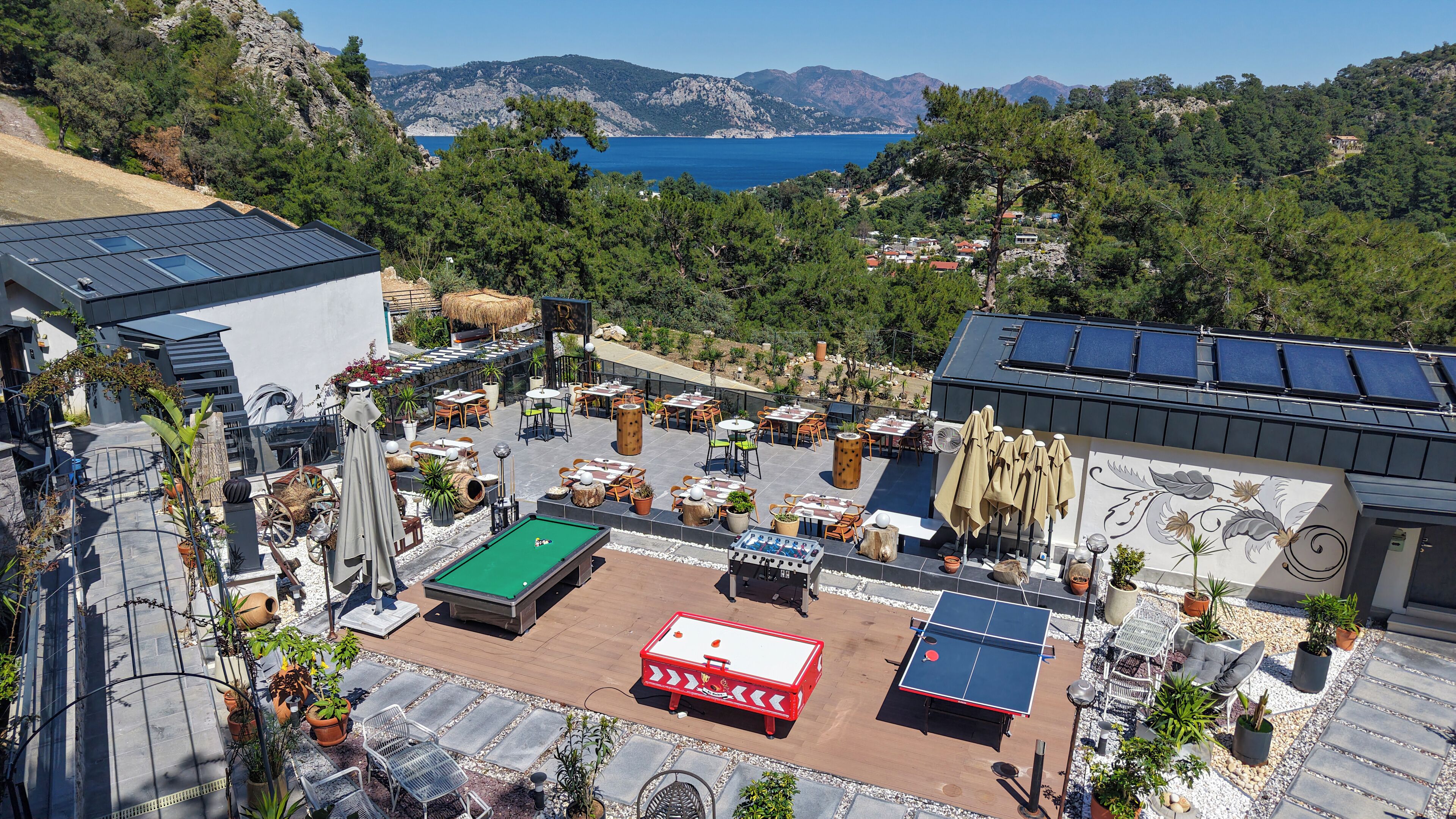 A rooftop terrace with a pool table, ping pong table, and outdoor seating area overlooking a scenic coastal landscape.