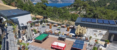 A rooftop terrace with a pool table, ping pong table, and outdoor seating area overlooking a scenic coastal landscape.