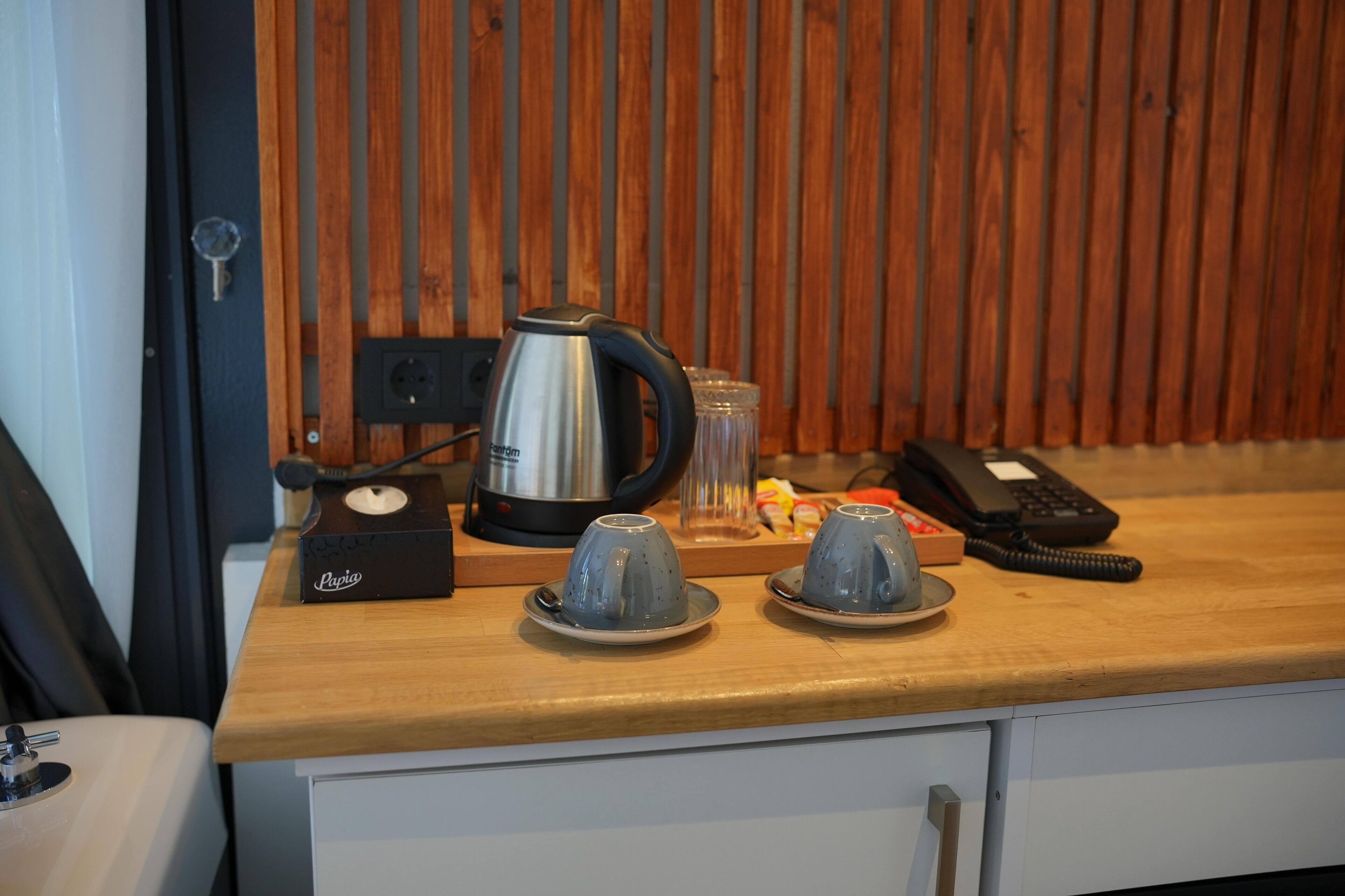 A kitchen counter with a coffee maker, two cups, a phone, and a kettle.