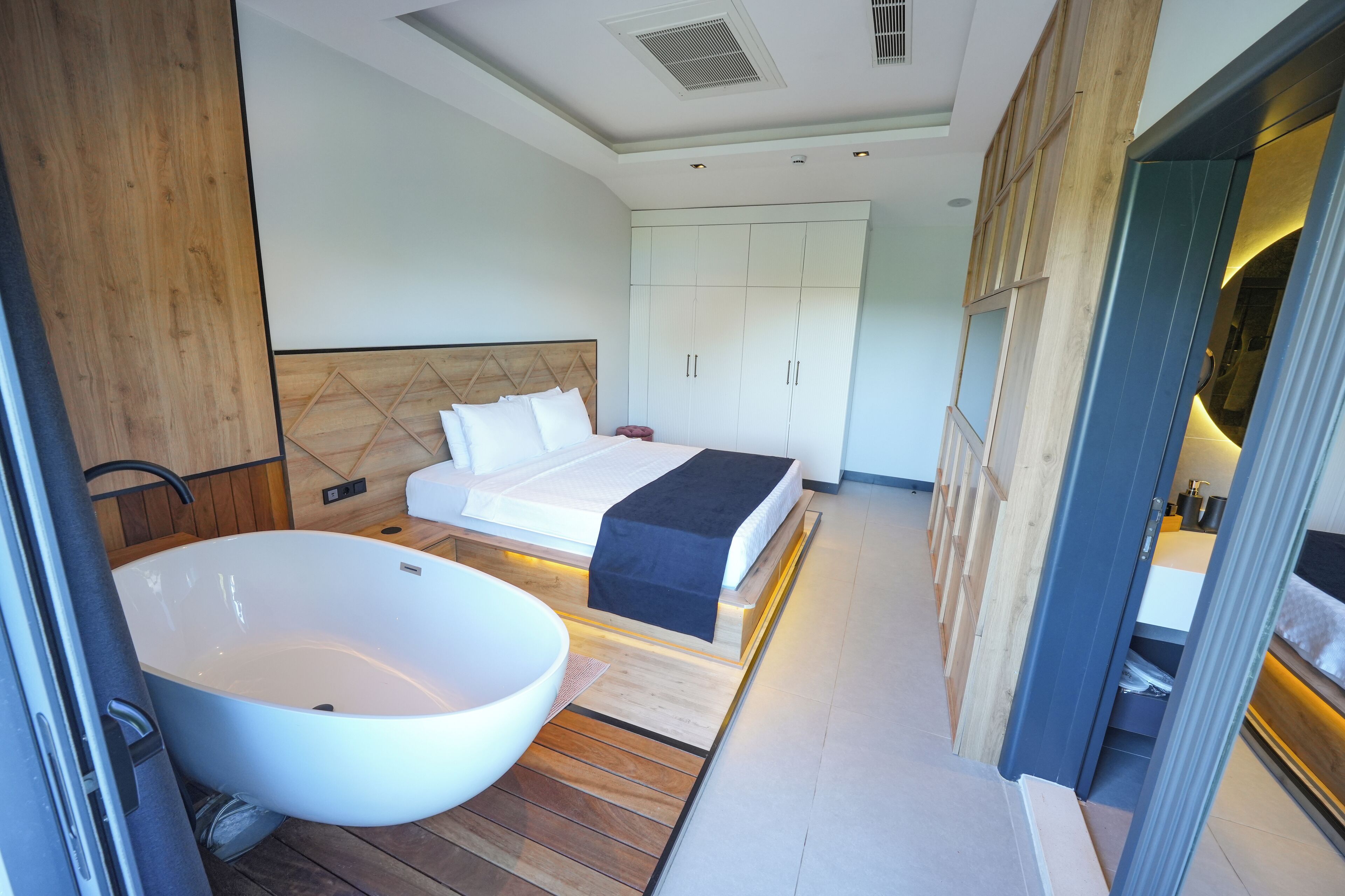 A modern bedroom with a large bathtub, a bed with a wooden headboard, and built-in wardrobes.