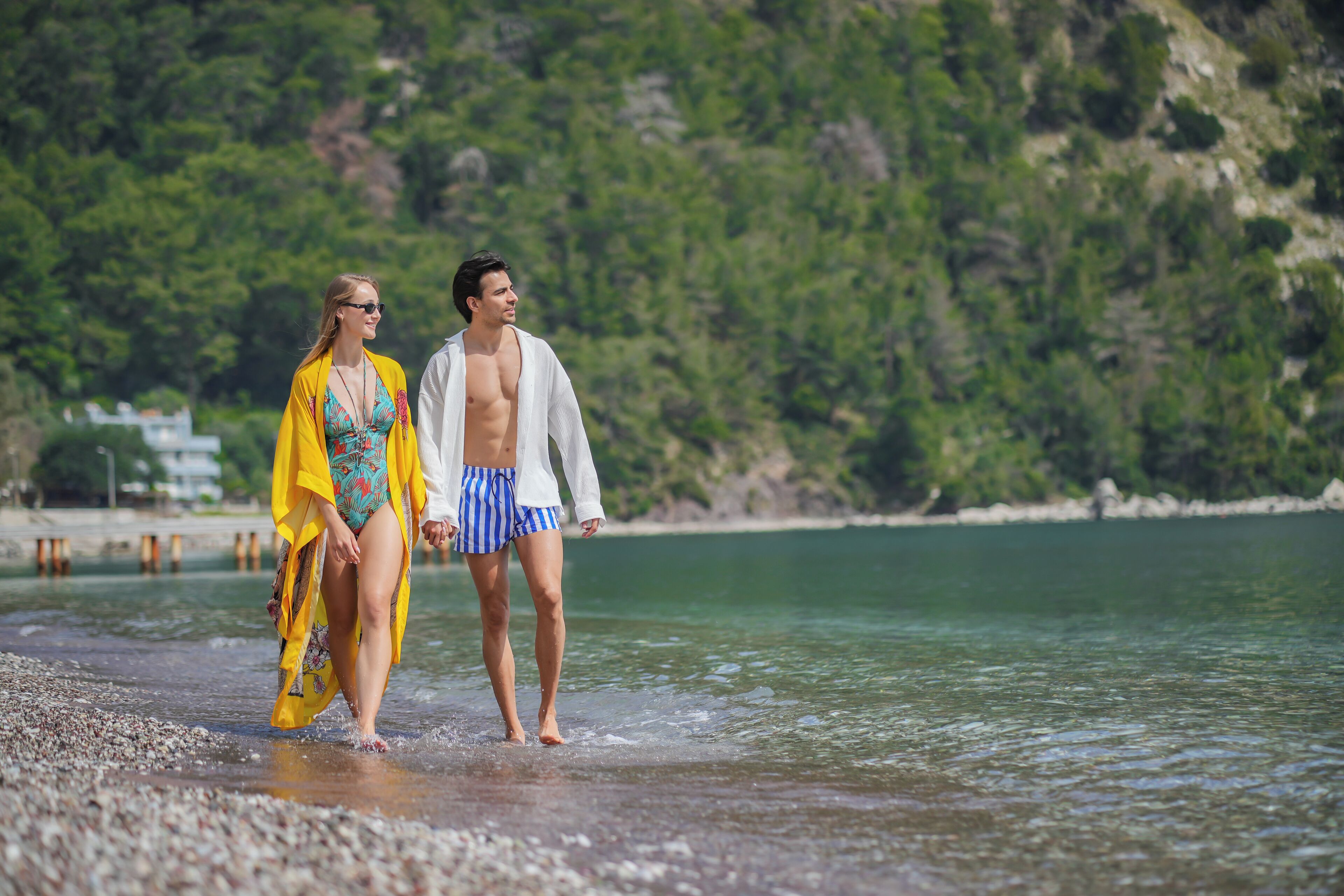 A couple walking along a pebbled beach with a rocky cliff and greenery in the background.