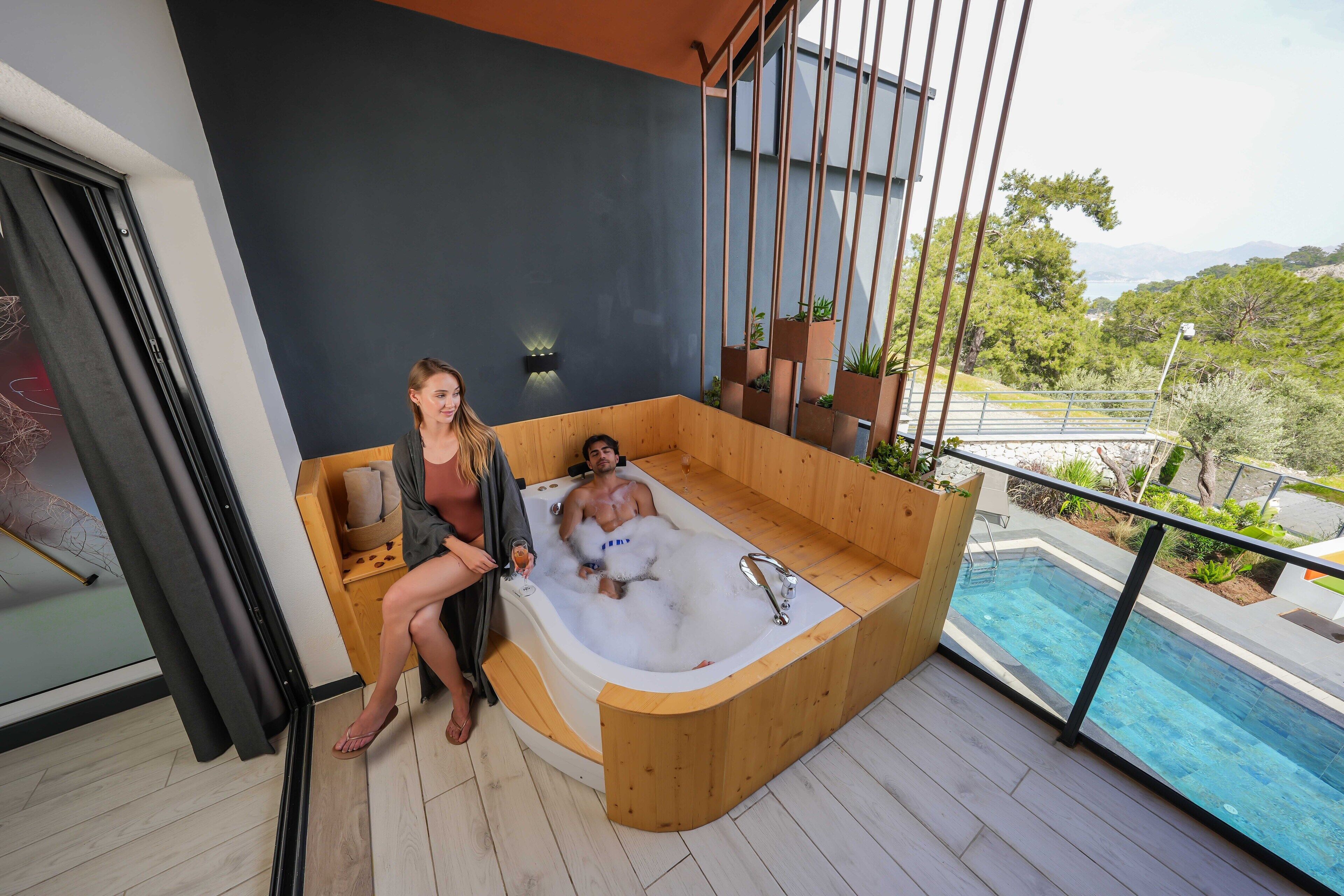 A modern outdoor hot tub with a wooden bench, a glass door, and a view of a pool and greenery.