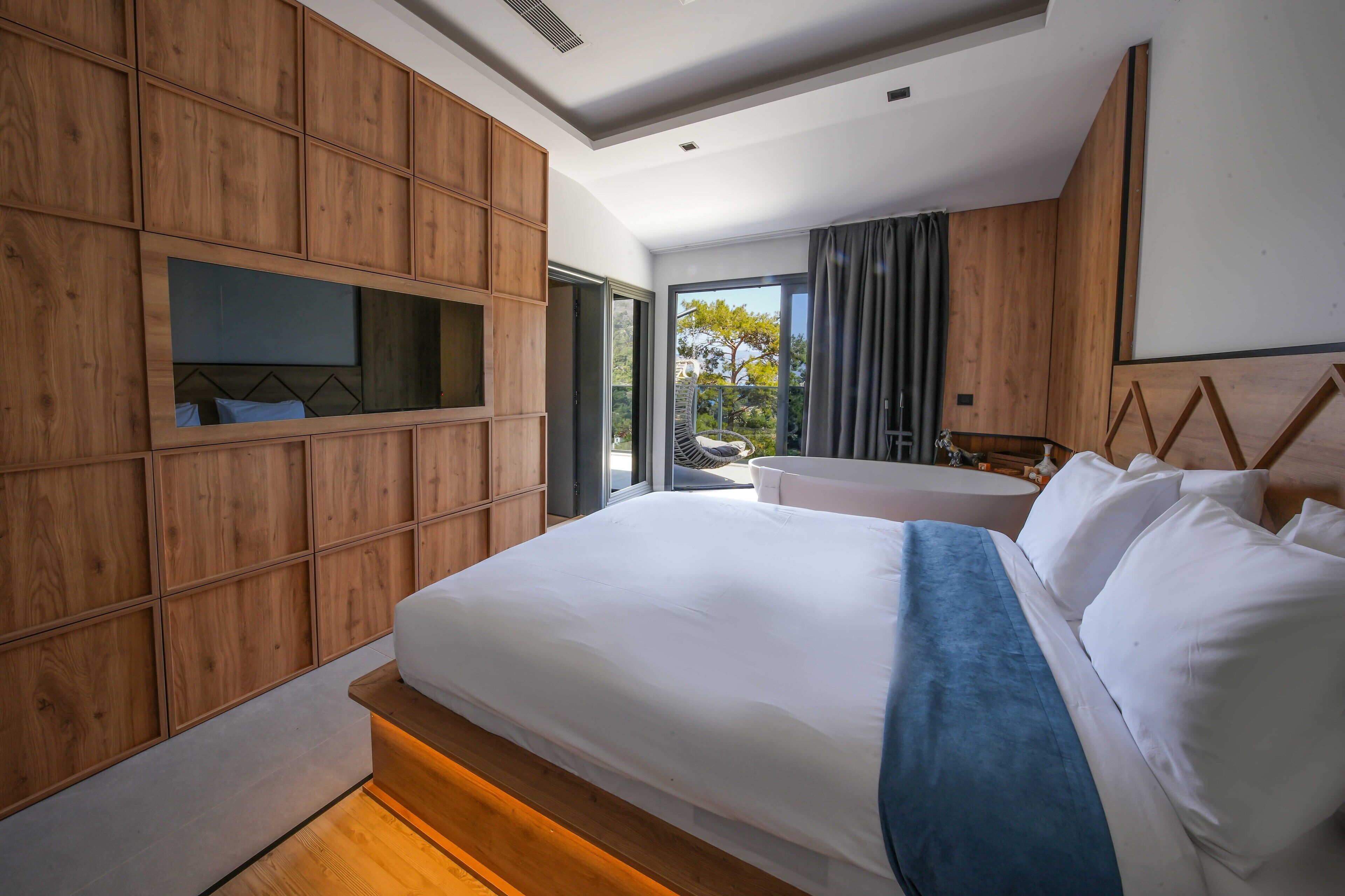 A modern bedroom with a large bed, wooden panel walls, a built-in TV, and a view of the outdoors.