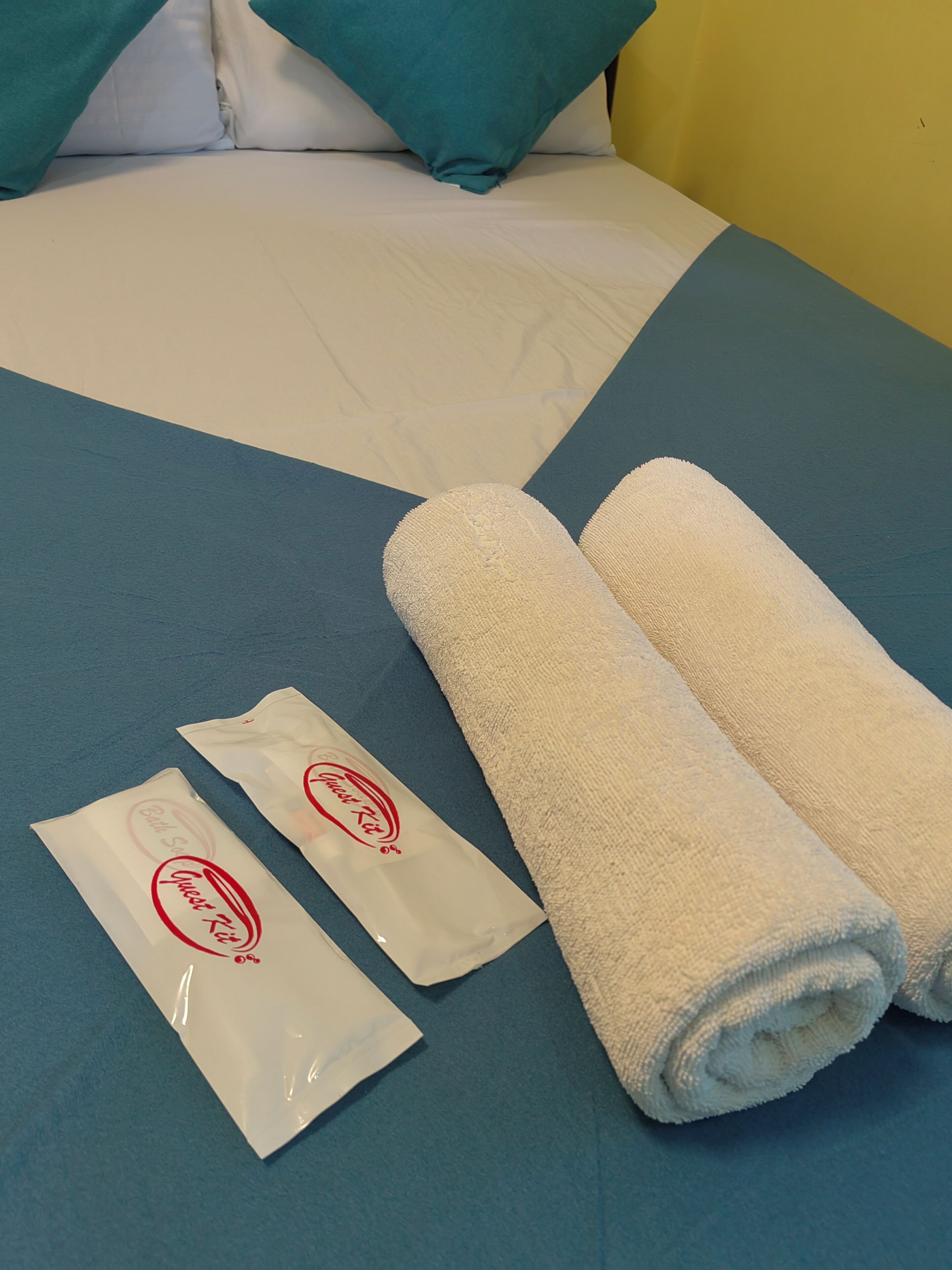 A bed with white and teal bedding, two rolled white towels, and two white bags with a logo on a blue bedspread.