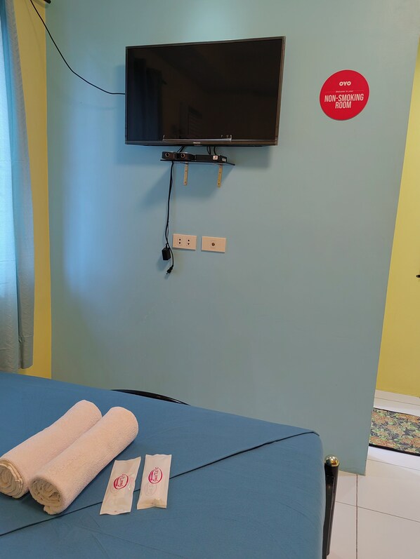 Habitación de hotel con un televisor de pantalla plana montado en la pared, una cama con colchón azul, dos toallas enrolladas y un cartel de no fumar.