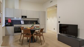 Una cocina moderna con armarios blancos, encimera negra y un comedor con una mesa redonda y cuatro sillas. Un televisor de pantalla plana está montado sobre un pedestal negro.