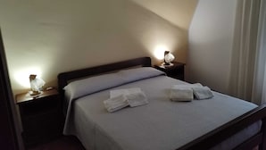 Un lit bien fait, recouvert d’une couvre-lit grise et agrémenté de deux oreillers, est placé de part et d’autre de deux tables de chevet sur lesquelles reposent des lampes. La pièce est simple, avec des murs unis et une fenêtre ornée de rideaux.