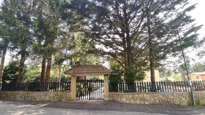 Une entrée fermée par un portail en métal et bordée d’un mur de pierre, le tout étant entouré de grands arbres.