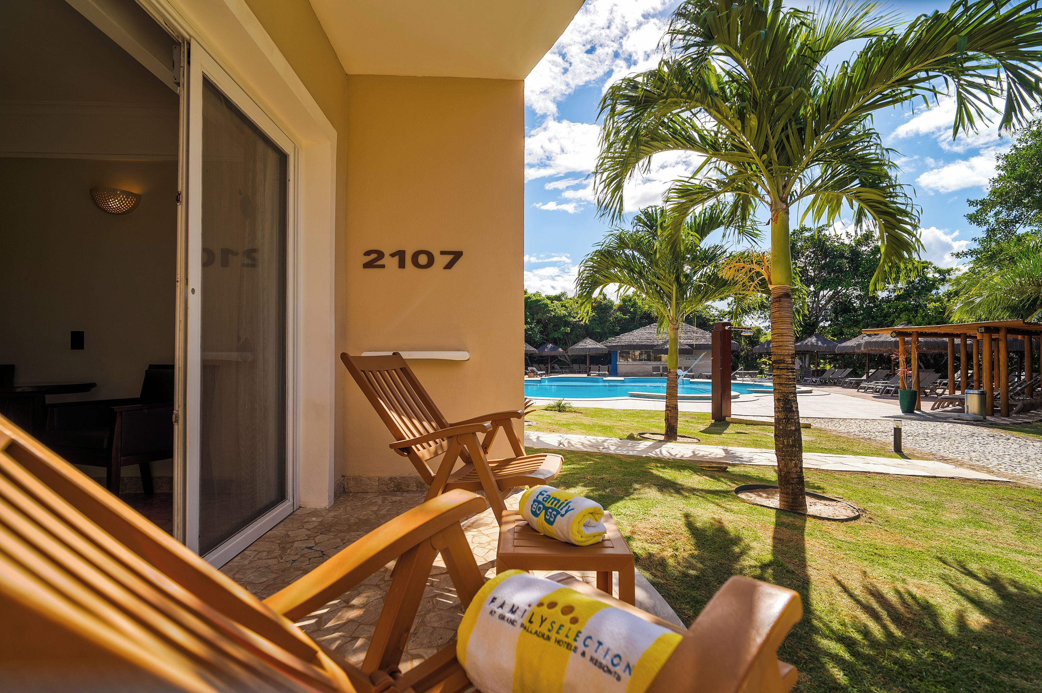 Quarto de hotel com porta de vidro deslizante, piscina e área externa com palmeira.