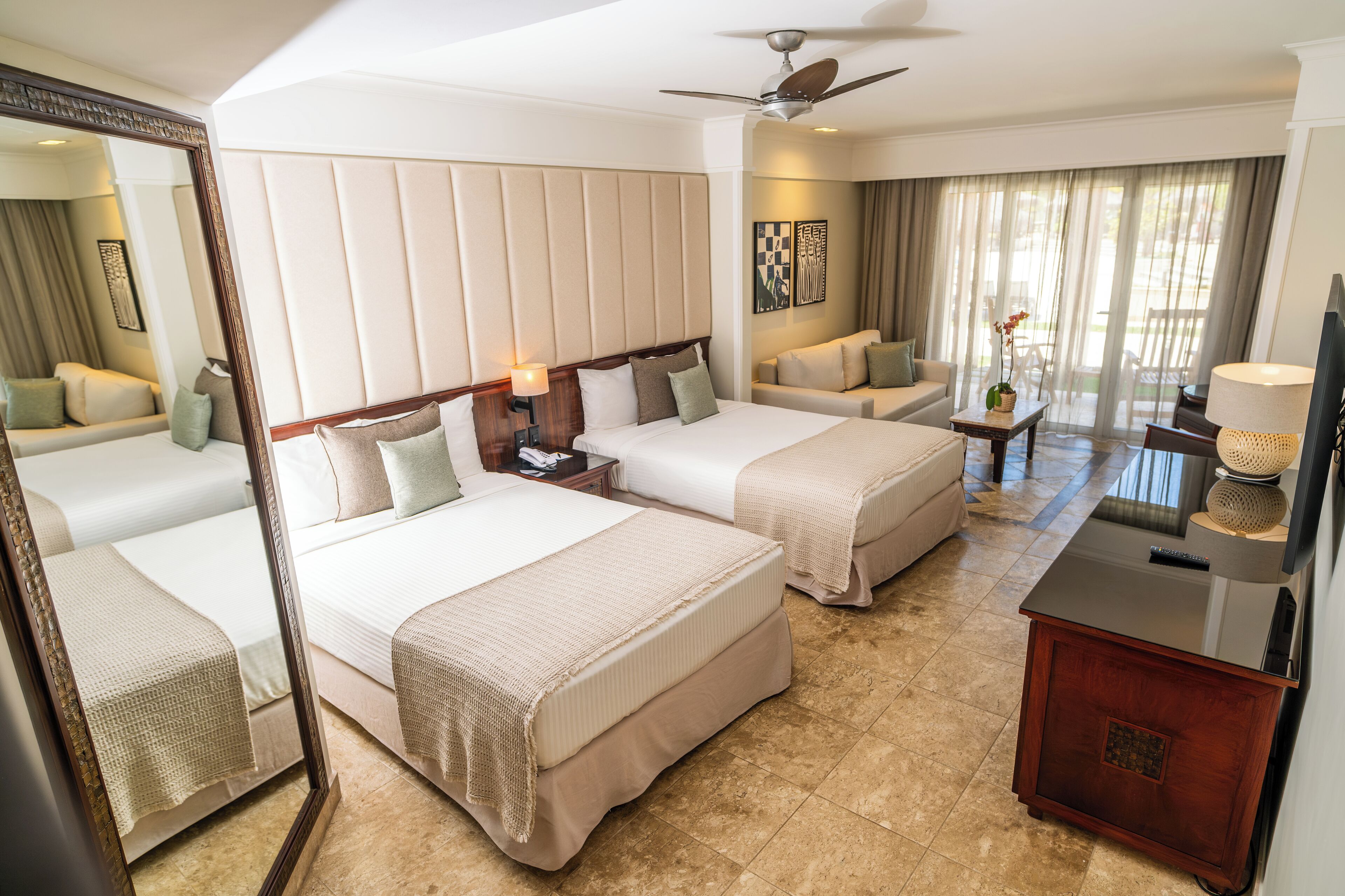 Quarto de hotel com duas camas, uma escrivaninha, uma cadeira, televisão e ventoinha de teto.