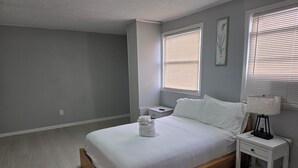 Una camera da letto con un letto in legno, biancheria da letto bianca, un comodino con una lampada e due finestre con persiane.