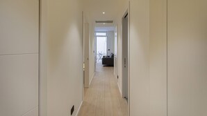 A narrow hallway with light-colored walls and a wooden floor, leading to a room with a sofa and a table.