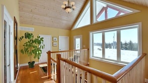 A spacious hallway with wooden floors, a staircase with a wooden railing, and large windows offering a view of the outdoors.