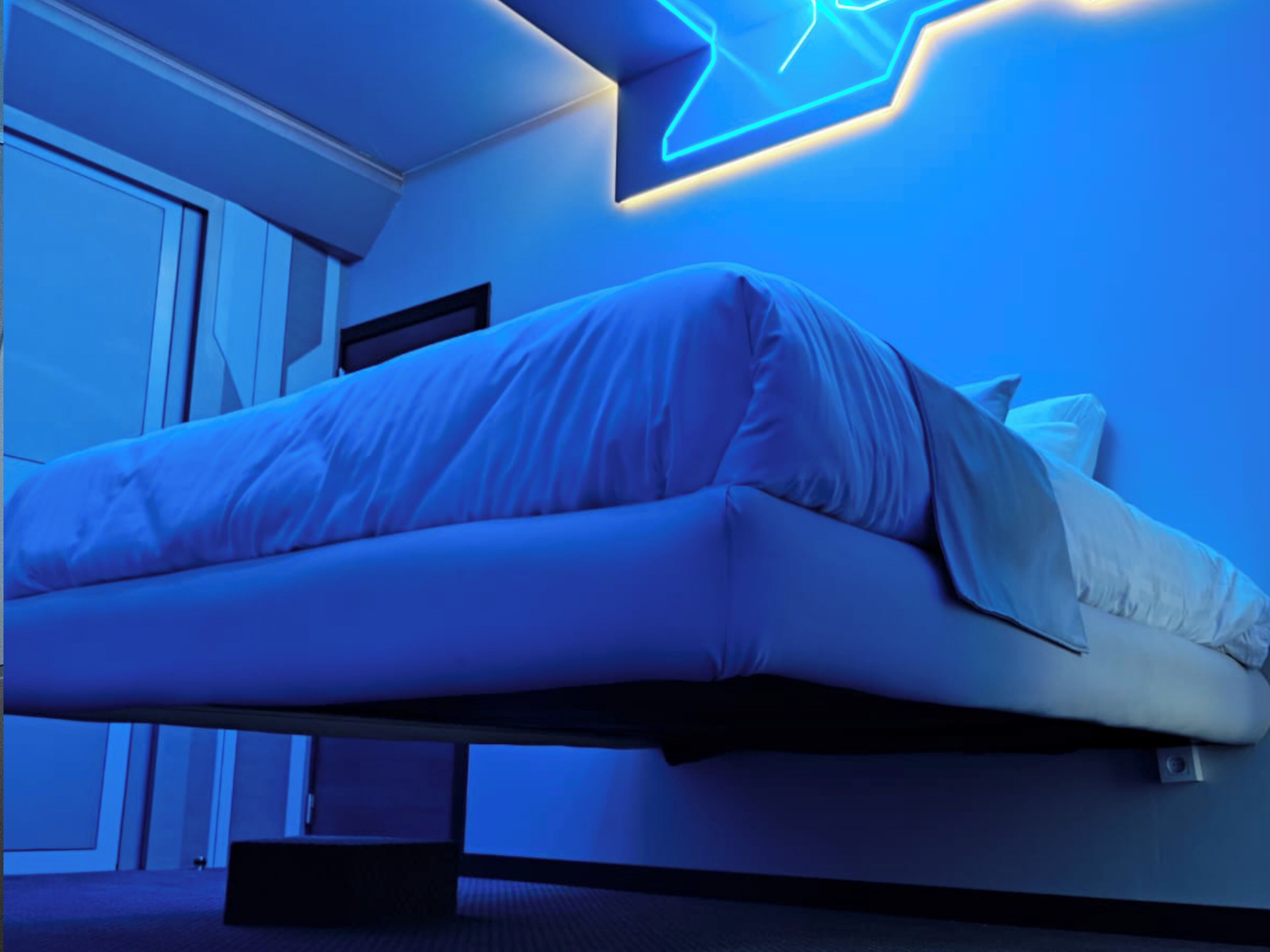 A bed in a room with a blue neon sign on the wall.