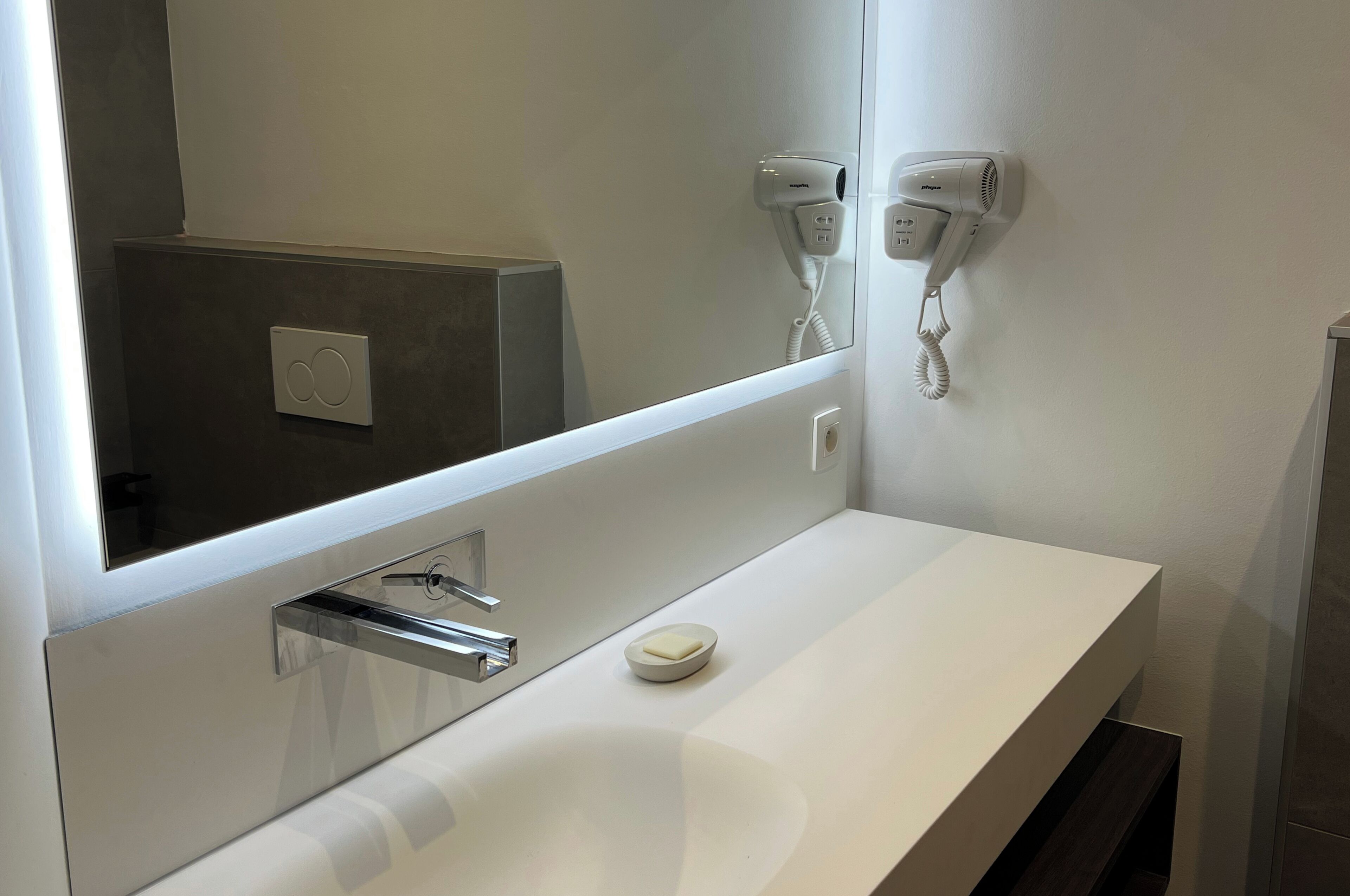 Een moderne badkamer met een witte wastafel, een spiegel, een haardroger en een zeepbakje.