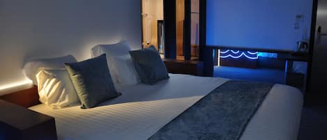 A neatly made bed with pillows and a blanket, a bedside table with a lamp, and a view of a wooden room through a doorway.