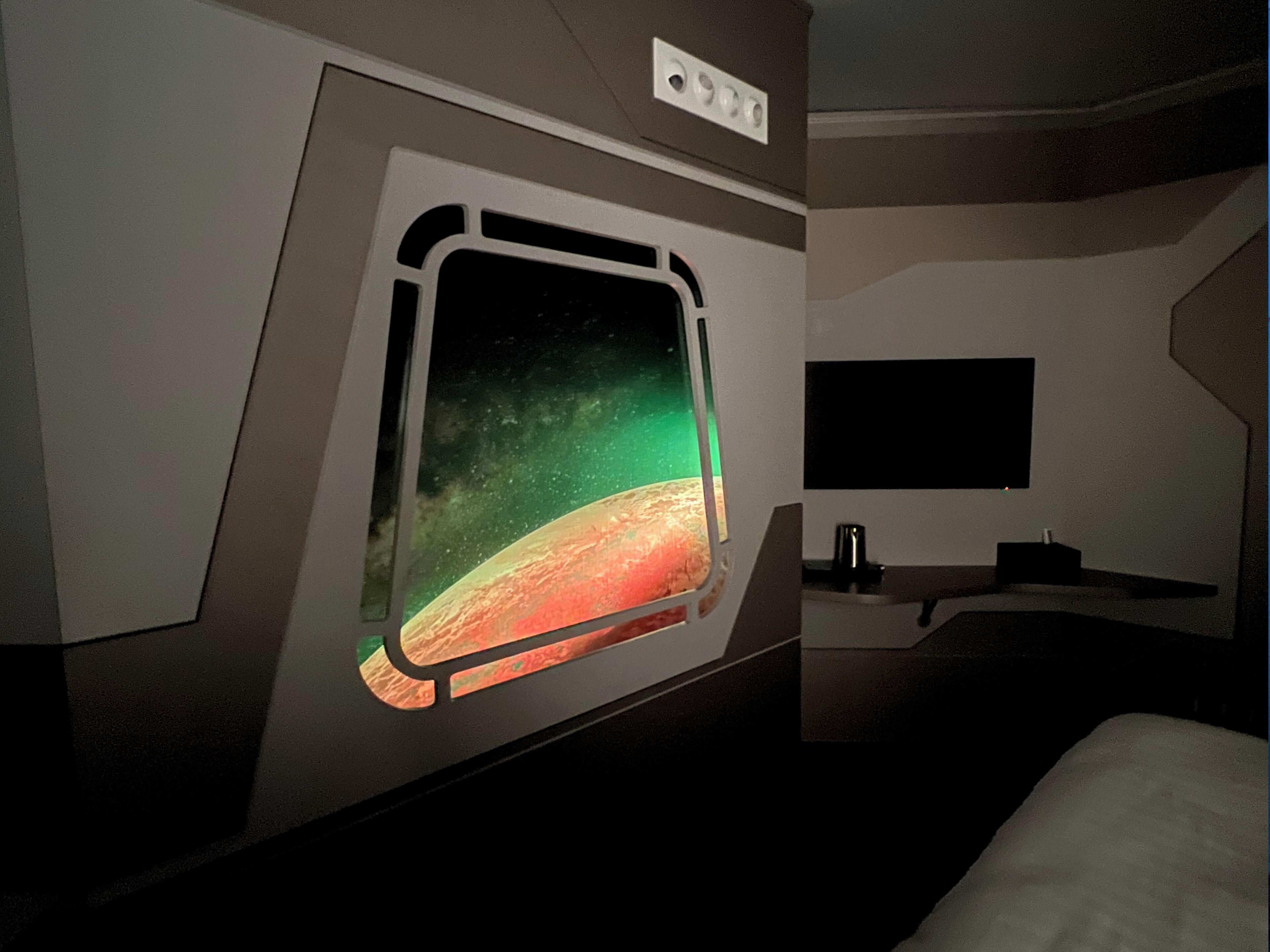 Una habitación con un ventanal grande que muestra una escena espacial, un televisor de pantalla plana montado en la pared y una mesita con una taza.