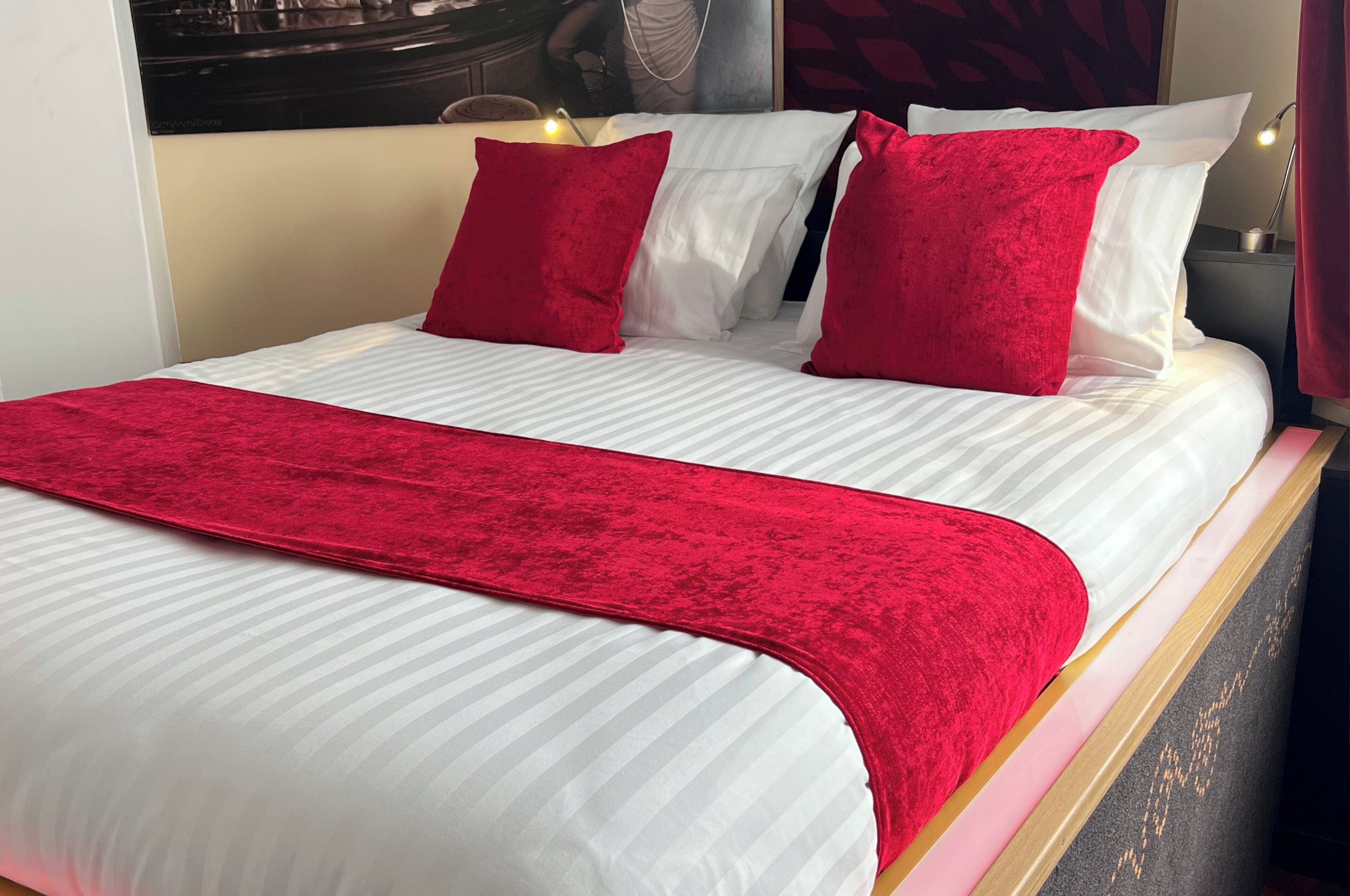 Een net opgemaakt bed met wit en rood beddengoed, een rode plaid en een hoofdbord met spiegel en verlichtingsarmatuur.